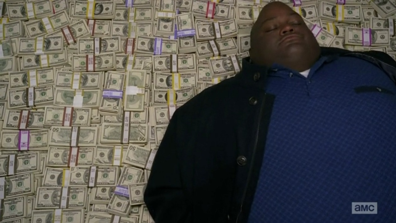 A still from the show 'Breaking Bad' showing the character Huell rolling atop an enormous pile of money.